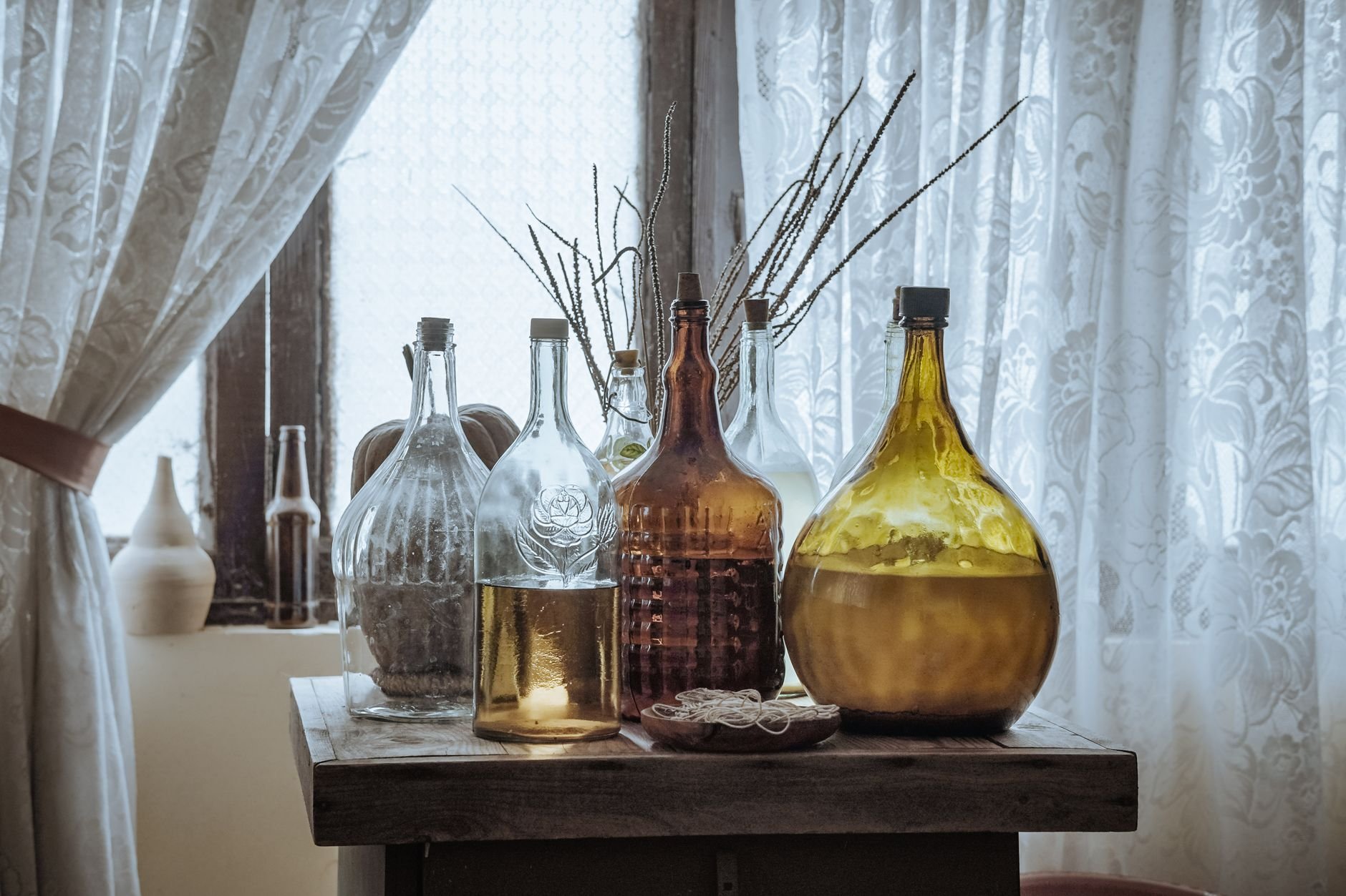 Aesthetic display of various vintage glass bottles on a rustic wooden table by a window with lace curtains.